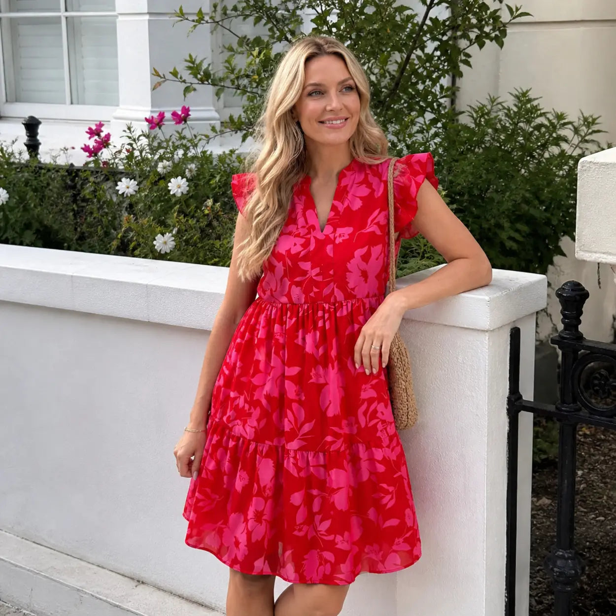 Woman in a red floral dress standing outdoors with greenery and flowers in the background